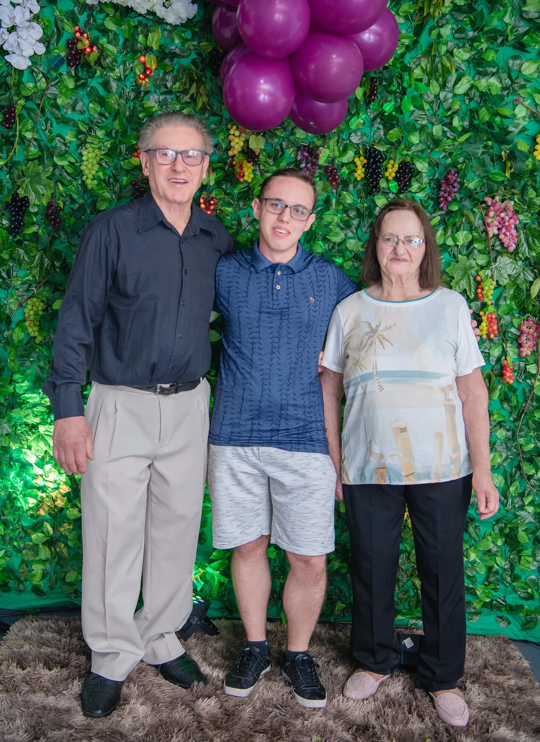 Um homem mais velho de óculos e camisa preta abraça um jovem de óculos e camisa polo azul, que está abraçando uma mulher mais velha de óculos e blusa branca com estampa de praia. Os três estão em pé na frente de uma parede verde decorada com folhas, uvas de plástico e um cacho de balões roxos no canto superior direito.