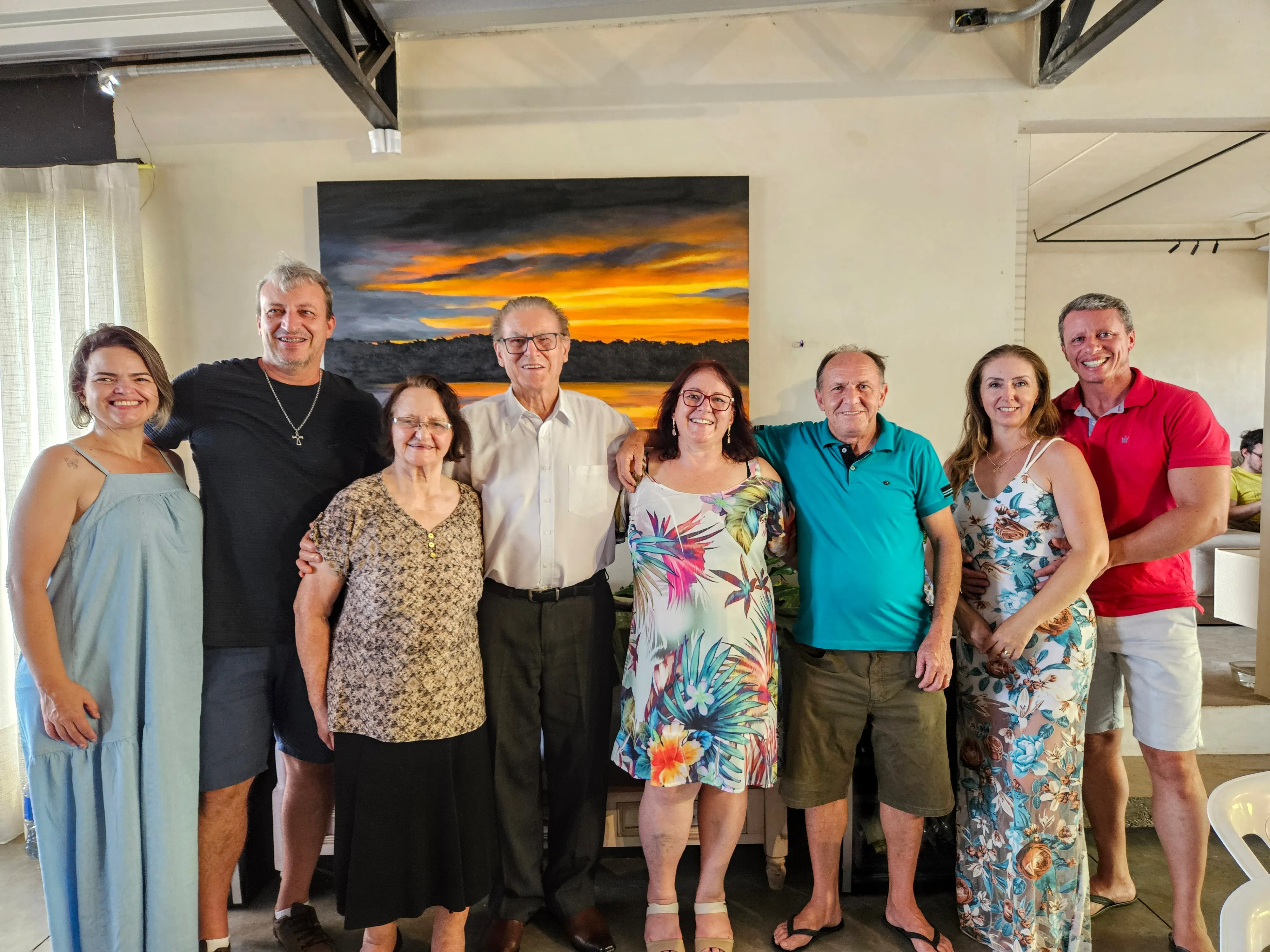 Um grupo de sete adultos, três homens e quatro mulheres, está em pé e sorrindo para a câmera dentro de casa. Atrás deles, há uma grande pintura de uma paisagem ao pôr do sol.