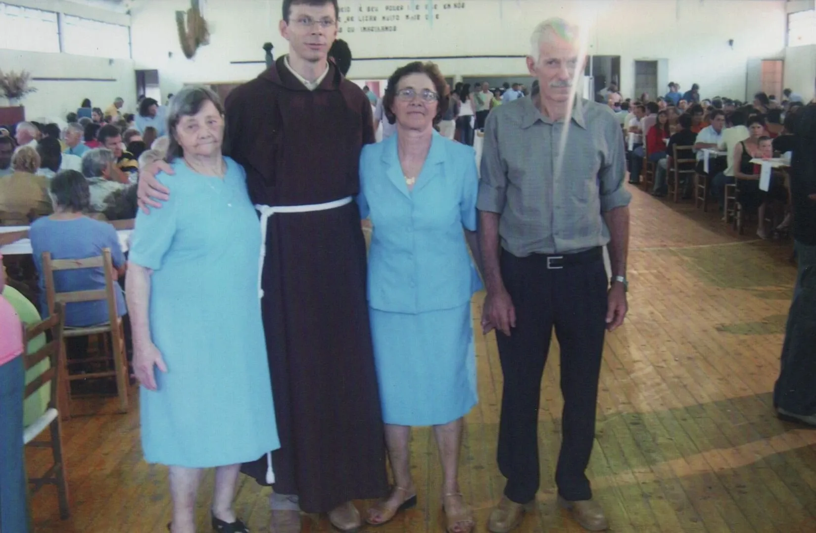 Quatro adultos, dois homens e duas mulheres, em pé lado a lado num grande salão de madeira com muitas pessoas sentadas em mesas ao fundo.