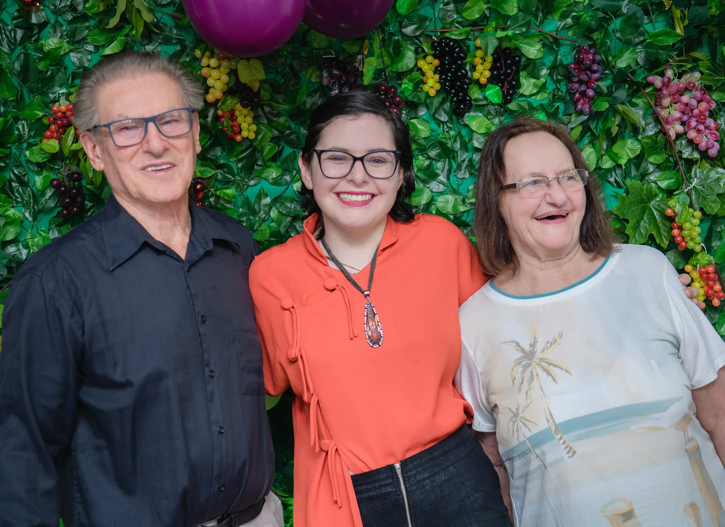 Três pessoas, um homem e duas mulheres, estão sorrindo para a câmera em frente a uma parede coberta de folhas verdes e cachos de uvas.