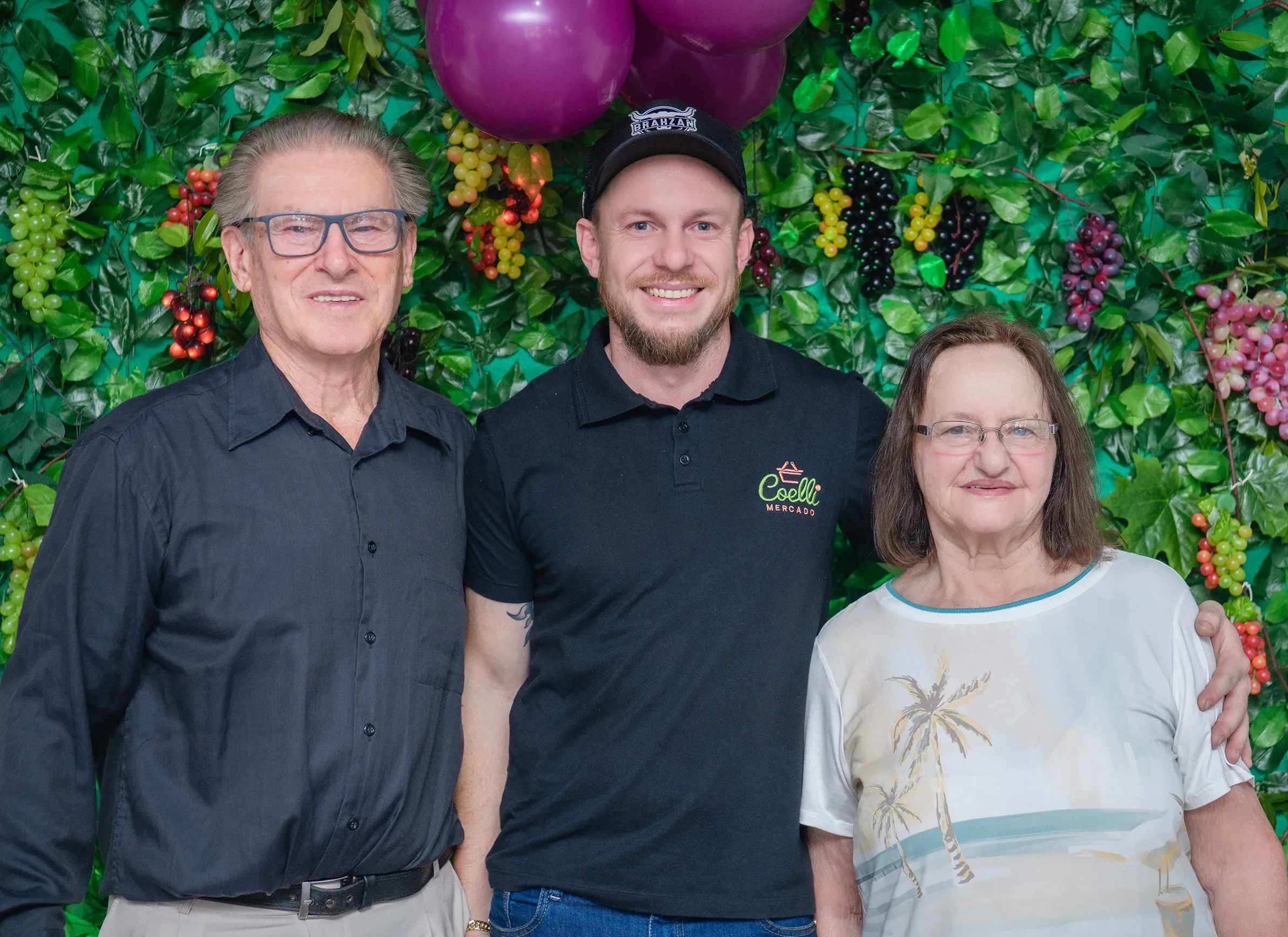 Um homem mais velho, um homem de meia-idade e uma mulher mais velha sorriem para a câmera em frente a uma parede coberta de folhas verdes e cachos de uvas artificiais; o homem de meia-idade veste uma camiseta preta com um logotipo branco.