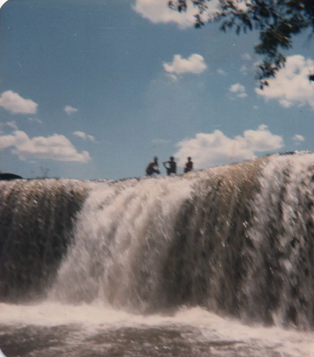 Três pessoas vistas de trás no topo de uma cachoeira em cascata sob um céu parcialmente nublado, com folhas escuras no canto superior direito. (Original)
