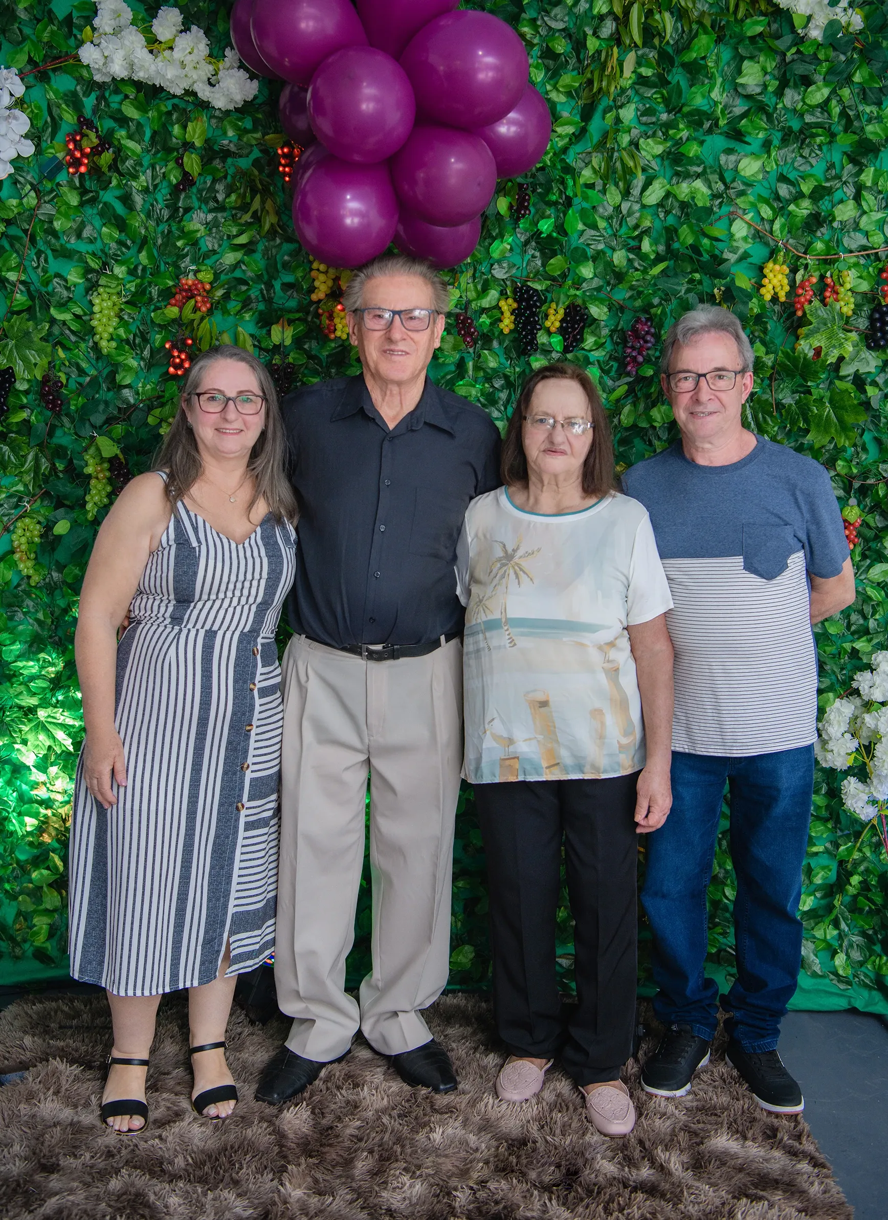 Um grupo de quatro pessoas, dois homens e duas mulheres, estão de pé e sorrindo para a câmera, com um fundo verde de hera falsa adornado com frutas e balões roxos.
