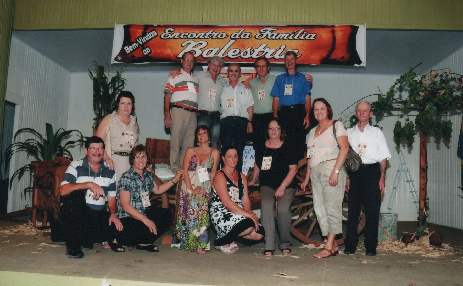 Um grupo de quinze adultos, dez mulheres e cinco homens, posam para uma fotografia em um palco sob uma faixa que diz "Bem-vindos ao Encontro da Família Balestrin".