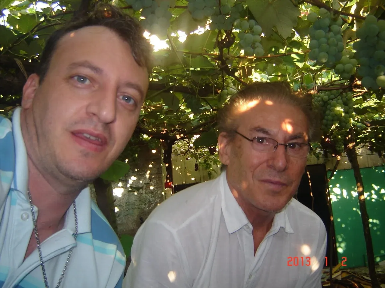 Dois homens posam para uma selfie ao ar livre, o da esquerda é um homem de meia-idade com cabelo castanho escuro e camisa pólo listrada azul e branca e um colar de corrente prata, olhando para a câmera e o da direita é um homem idoso com cabelo ruivo e uma camisa branca, olhando para a câmera com óculos.