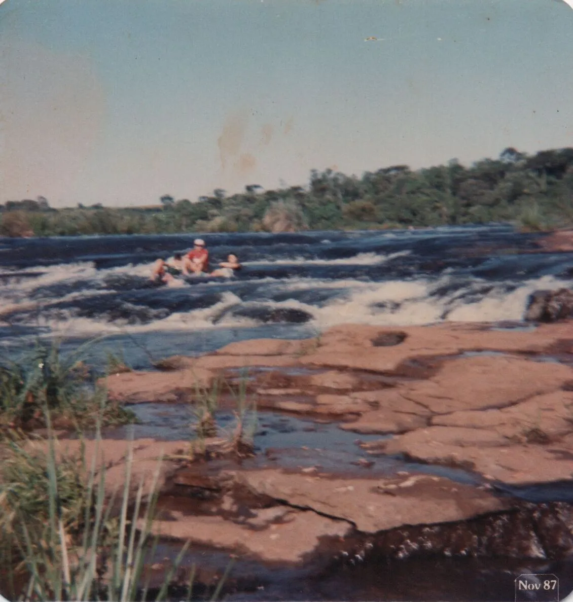Pessoas em um rio turbulento próximo a uma área rochosa com árvores ao fundo. (Original)