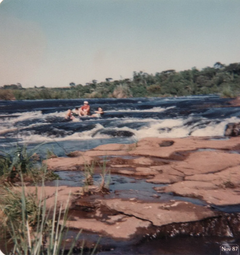 Pessoas em um rio turbulento próximo a uma área rochosa com árvores ao fundo.