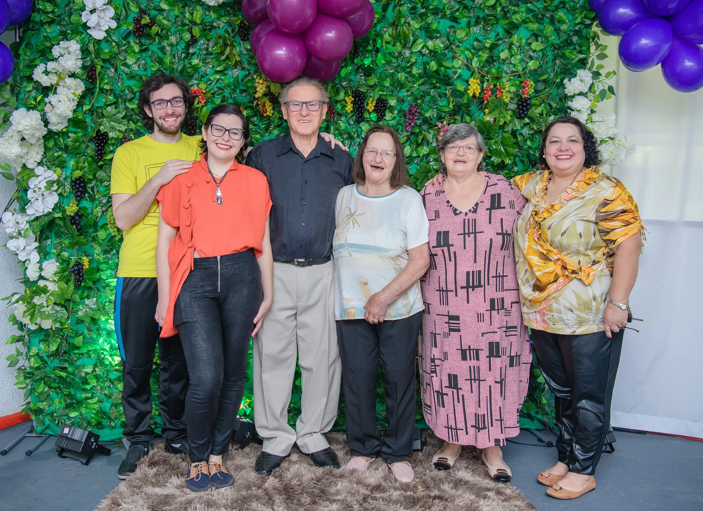 Um grupo de sete pessoas, incluindo um homem de camisa amarela, uma mulher de camisa laranja, um homem de camisa preta, duas mulheres mais velhas e duas mulheres de meia-idade, posando juntos na frente de uma parede coberta de folhas verdes com cachos de uvas roxas e flores brancas e roxas.