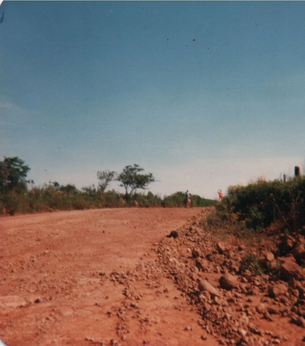 Uma estrada de terra marrom escura e empoeirada se estende para longe sob um céu azul claro, ladeada por árvores e arbustos verdes exuberantes em ambos os lados, com duas pessoas ao longe. (Original)
