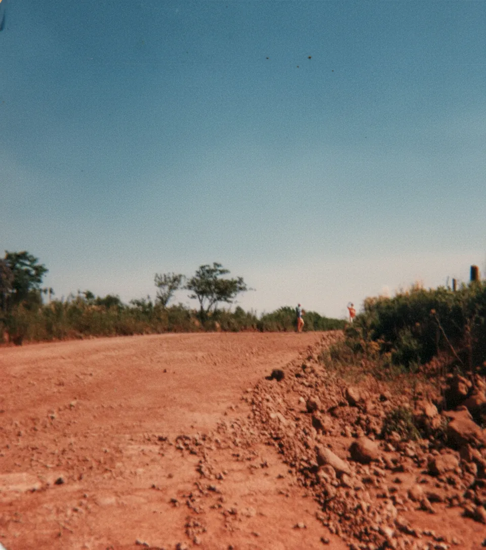 Uma estrada de terra marrom escura e empoeirada se estende para longe sob um céu azul claro, ladeada por árvores e arbustos verdes exuberantes em ambos os lados, com duas pessoas ao longe.