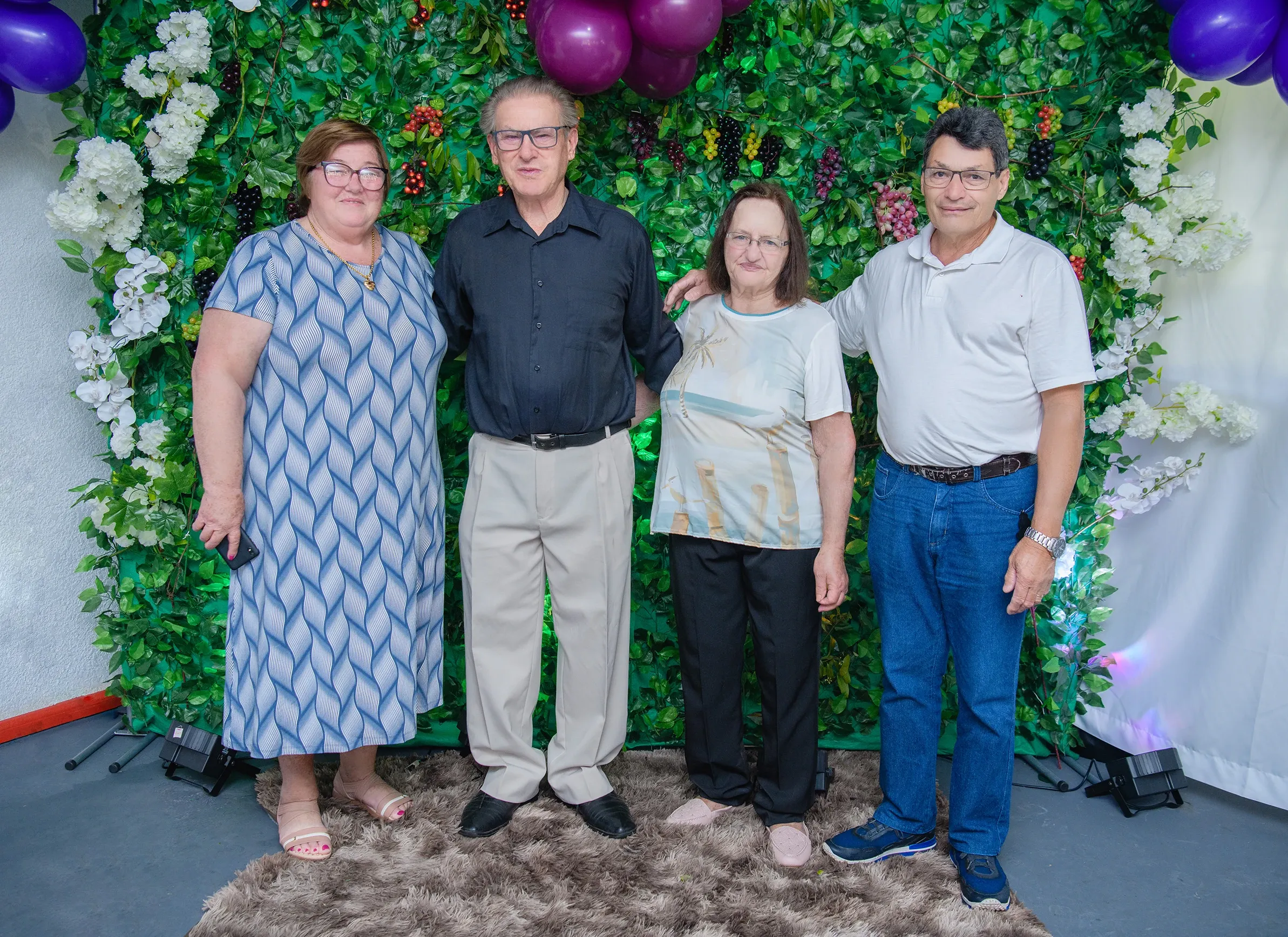 Quatro pessoas sorridentes, dois homens e duas mulheres, estão lado a lado em frente a uma parede decorada com folhagem verde, flores brancas e cachos de uva.