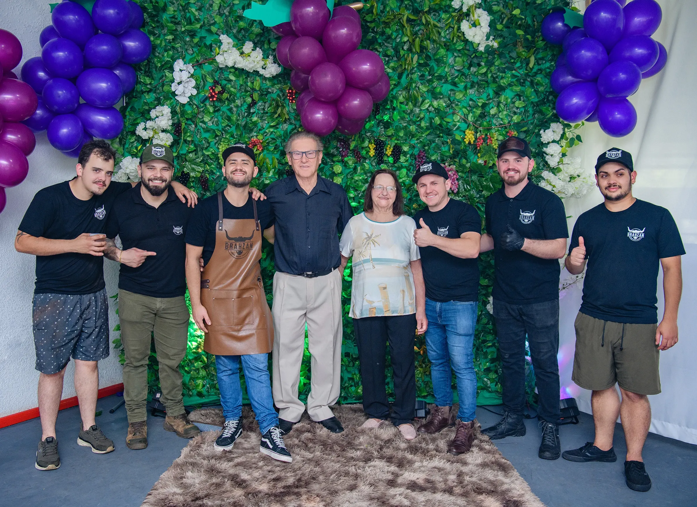 Um grupo de nove pessoas, a maioria homens jovens, está de pé na frente de uma parede coberta por folhas verdes e cachos de uvas roxas. Quatro homens à esquerda, todos sorrindo, usam camisetas pretas e bonés, um deles com um avental de couro. No centro, um homem e uma mulher mais velhos, também sorrindo, estão ladeados por dois homens à direita, que usam camisetas pretas e bonés. O homem do centro veste uma camisa preta de mangas compridas e calças cáqui, enquanto a mulher usa uma blusa clara estampada e calças escuras. Todos parecem felizes e à vontade. O chão é coberto por um tapete felpudo marrom.