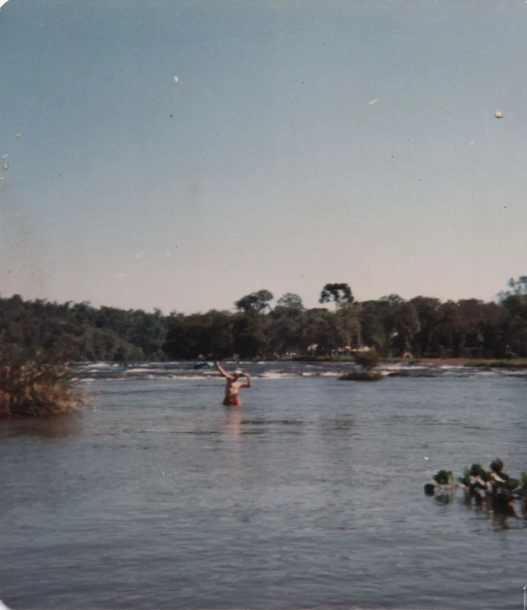 Um homem sem camisa, de bermuda vermelha, está parado até a cintura em um rio calmo, com os braços erguidos em uma pose de vitória; uma exuberante floresta verde-escura e um céu azul claro preenchem o fundo. (Original)