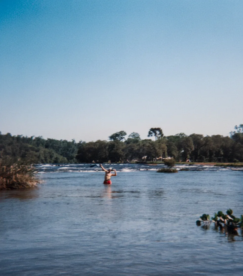 Um homem sem camisa, de bermuda vermelha, está parado até a cintura em um rio calmo, com os braços erguidos em uma pose de vitória; uma exuberante floresta verde-escura e um céu azul claro preenchem o fundo.