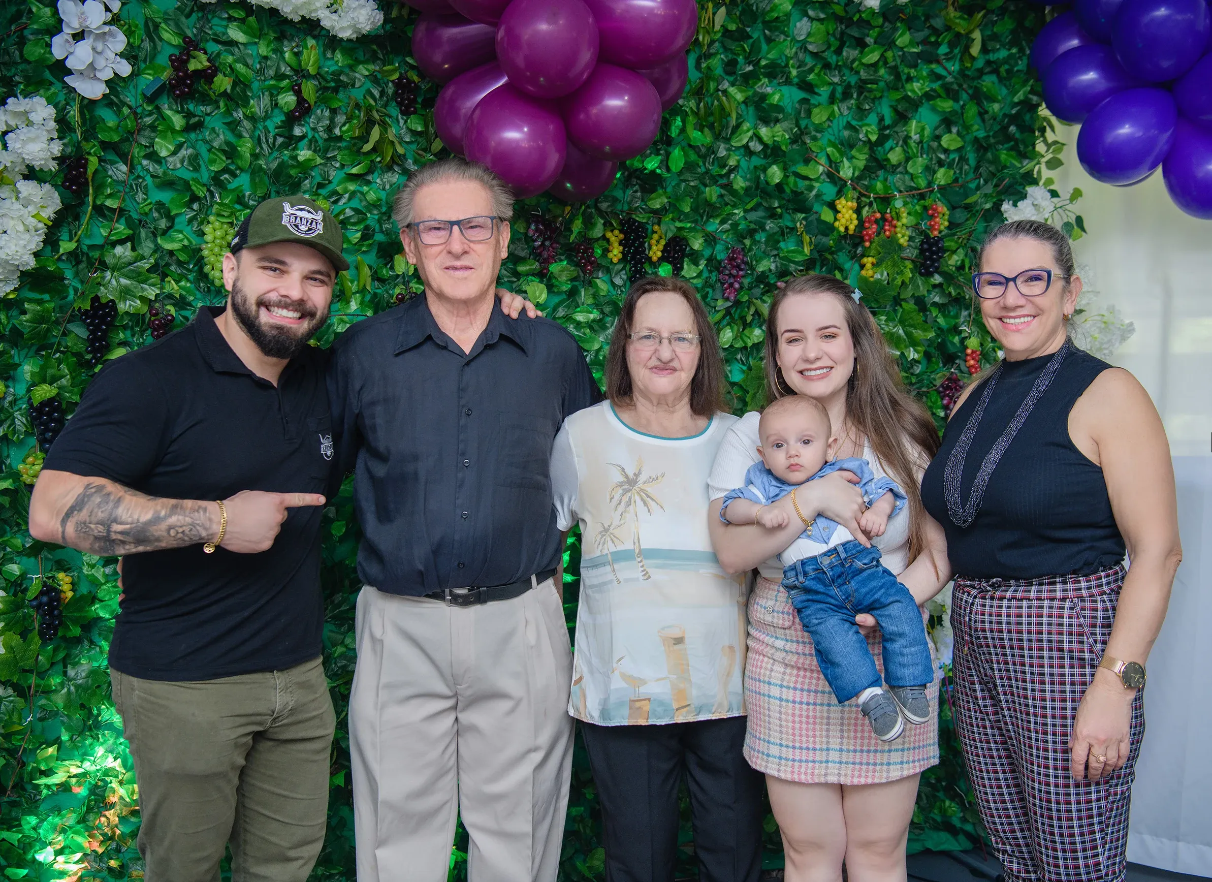 Uma família de três gerações posa para uma foto em frente a uma parede de folhagem com uvas roxas e balões.
