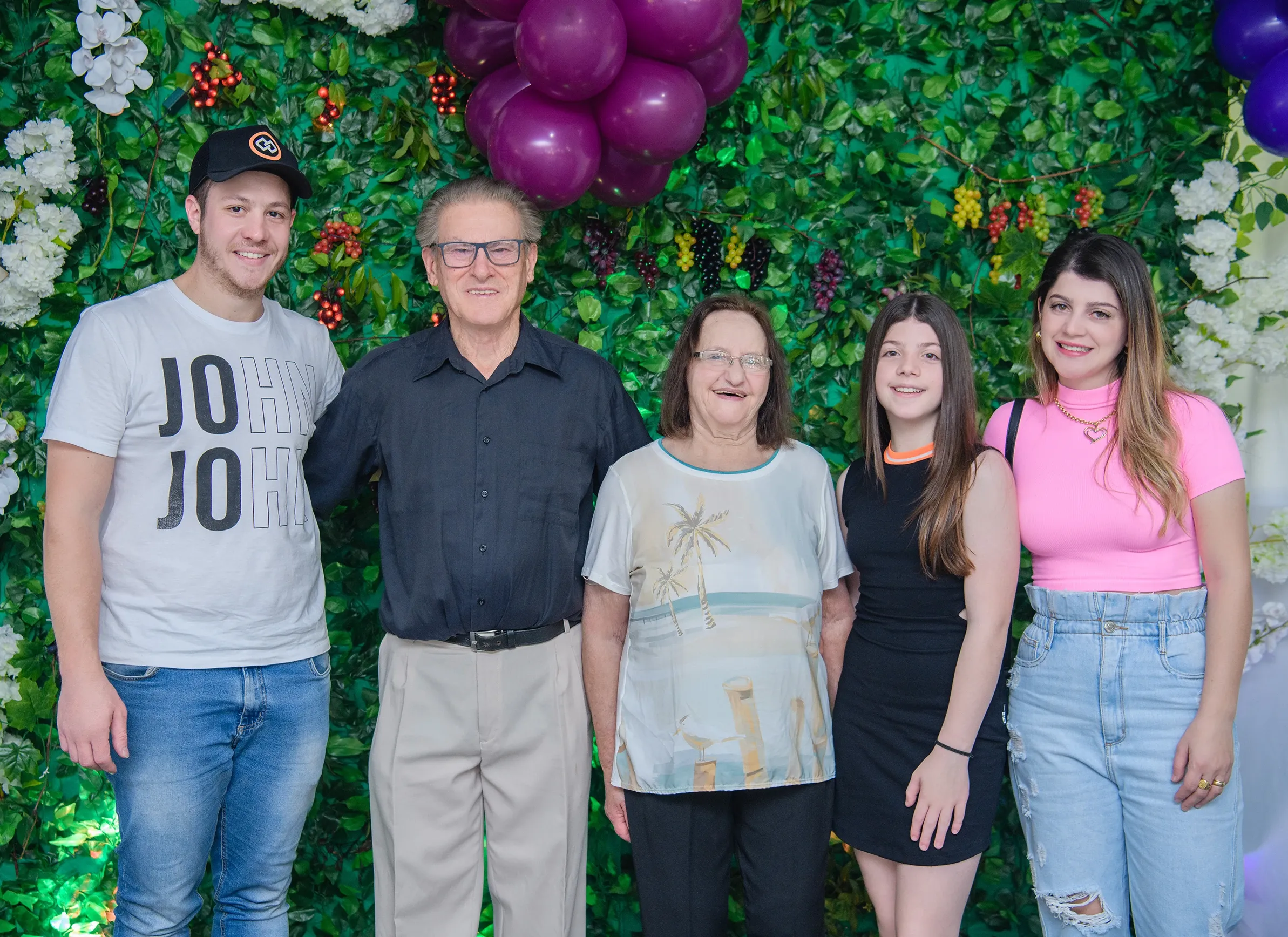 Um grupo de cinco pessoas está posando na frente de uma parede verde decorada com folhagens, flores brancas e roxas e cachos de uvas, com balões roxos visíveis no canto superior direito.