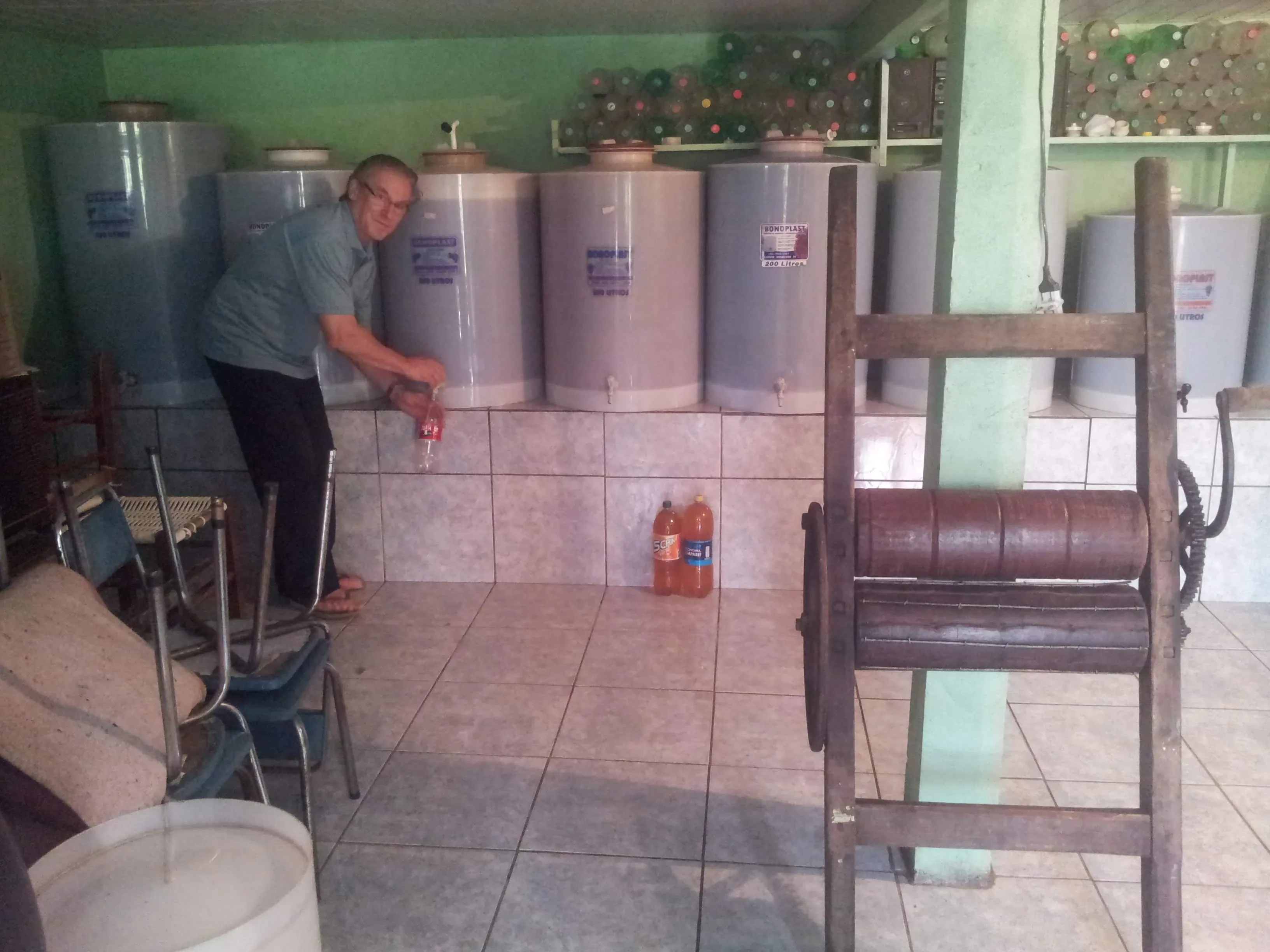 Um homem de óculos e camisa azul enche uma garrafa de Coca-Cola de um grande recipiente cilíndrico em um cômodo com paredes verde-claras e ladrilhos brancos; grandes tanques de armazenamento e várias garrafas plásticas estão ao fundo, e um espremedor de roupas de madeira está à direita.