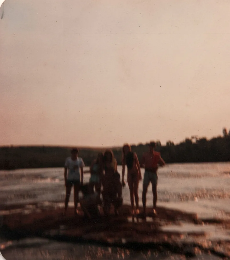Cinco pessoas de pé e uma agachada em um rochedo no meio de um rio largo, com um horizonte de árvores escuras sob um céu claro e cor de pêssego.