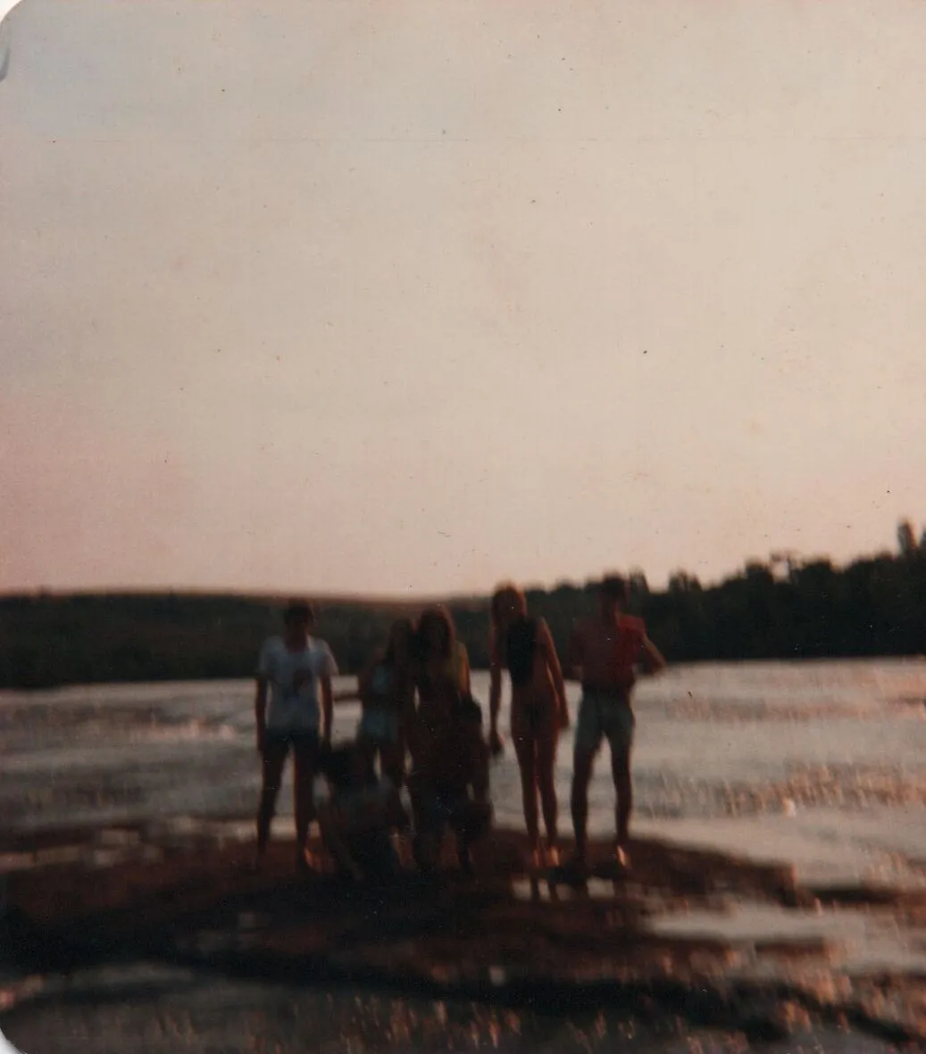 Cinco pessoas de pé e uma agachada em um rochedo no meio de um rio largo, com um horizonte de árvores escuras sob um céu claro e cor de pêssego. (Original)