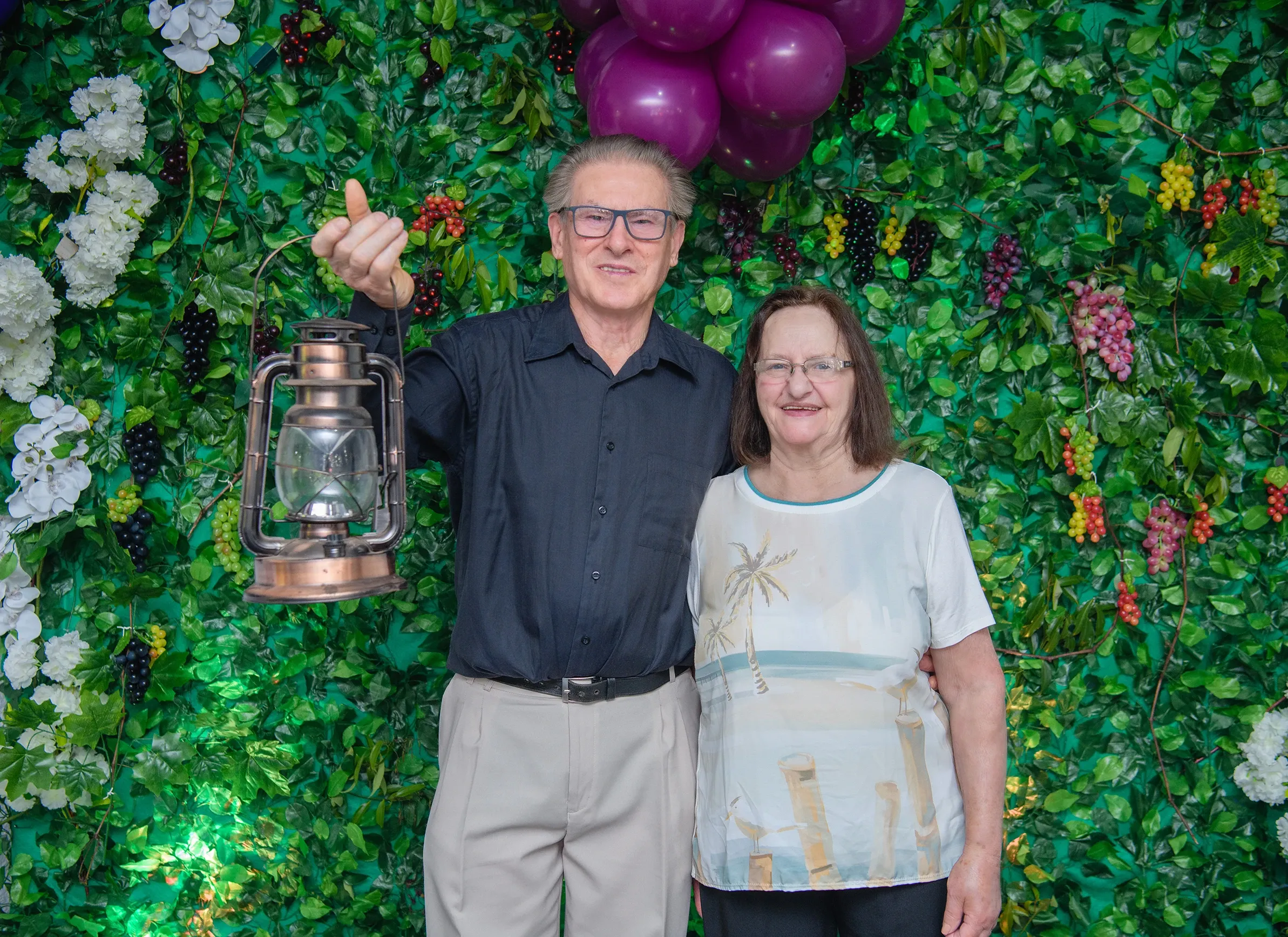 Um homem de camisa preta e calças bege segura uma lanterna antiga e uma mulher de camisa branca com estampa azul abraça o homem, ambos sorrindo na frente de uma parede verde com folhas e frutas.