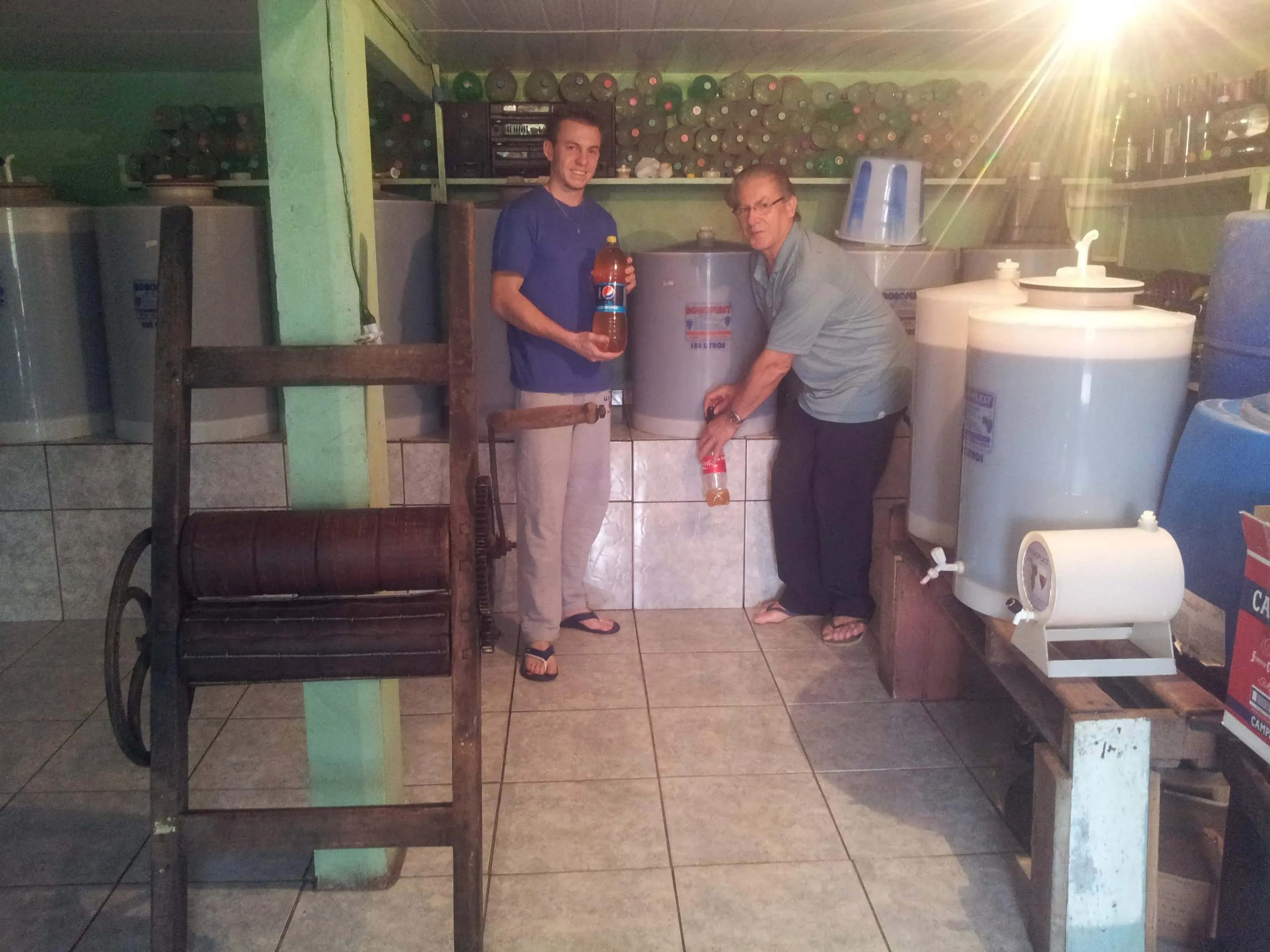 Dois homens, um jovem e outro idoso, engarrafando líquido de recipientes grandes em um ambiente de adega.