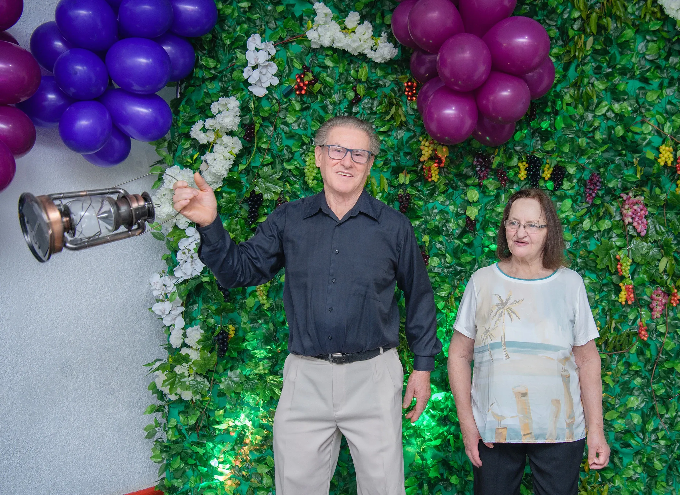 Um homem de óculos e camisa preta gesticula em direção a uma lanterna flutuante enquanto uma mulher de óculos e blusa branca com estampa de palmeiras está ao lado dele, tudo contra um fundo de parede de hera com balões roxos e brancos.