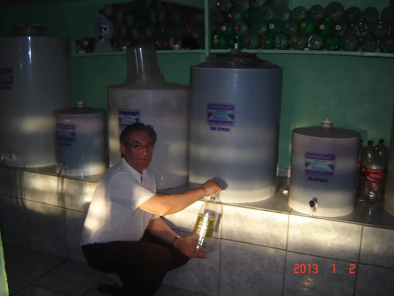 Homem agachado em uma sala de engarrafamento de sucos, segurando uma garrafa PET cheia de líquido amarelo ao lado de grandes tanques de suco.