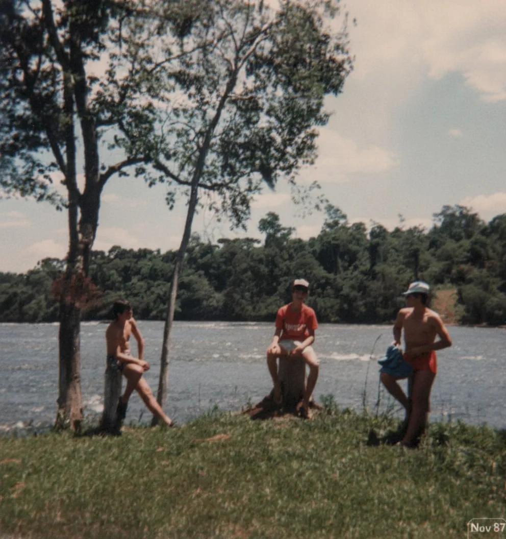 Três homens jovens em trajes de banho estão perto de uma grande árvore, com um em um toco e os outros dois de pé na grama ao lado de um rio largo e verdejante.