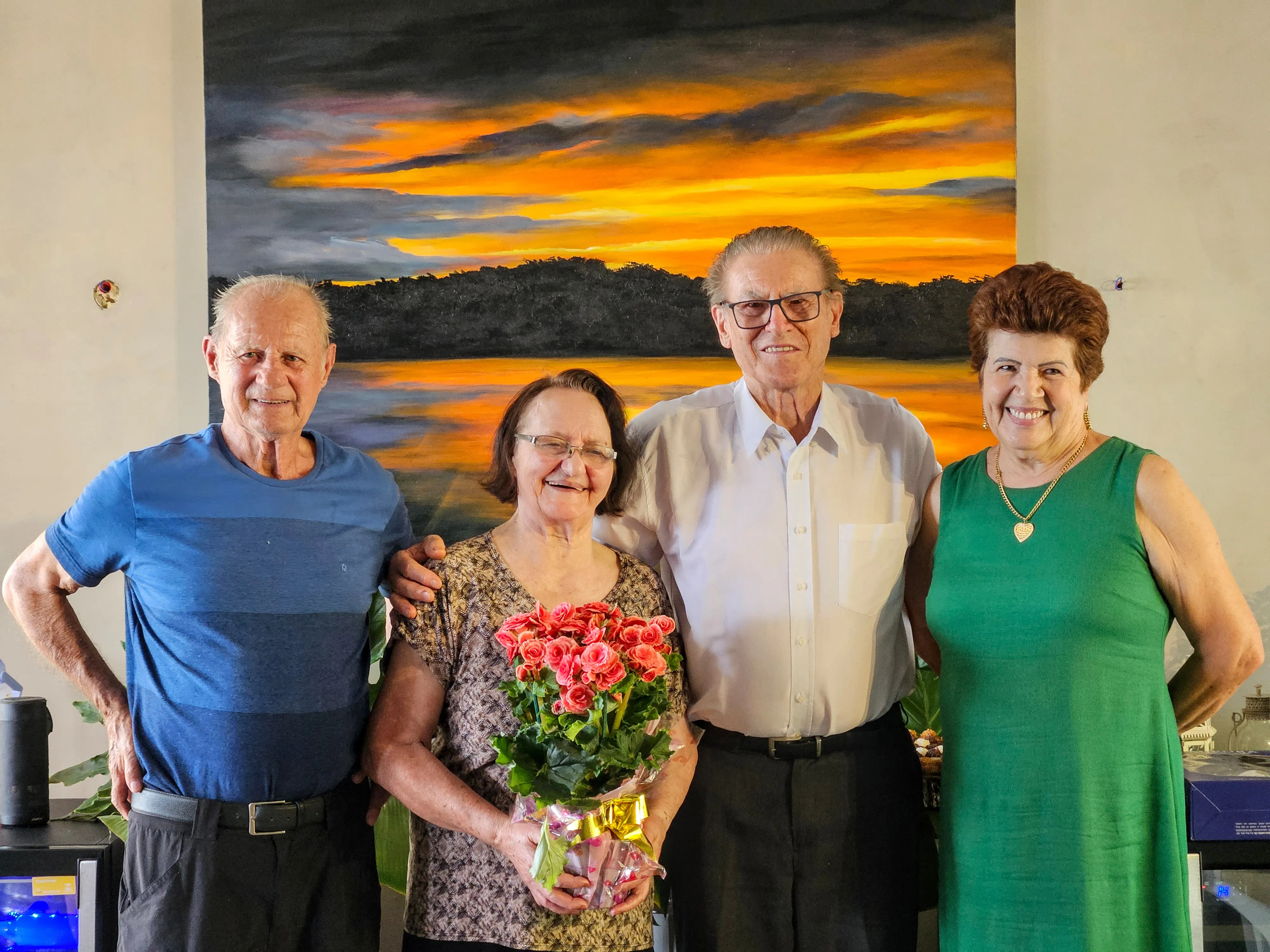 Quatro idosos sorriem para a câmera, com uma mulher no centro segurando um buquê de rosas vermelhas, à frente de uma pintura de pôr do sol.