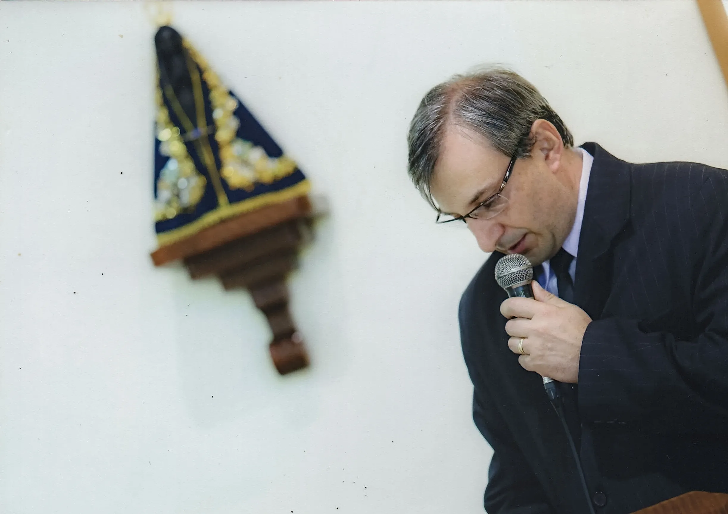 Um homem de terno e óculos segura um microfone e fala, com a cabeça baixa em oração, diante de uma estátua da Virgem Maria em uma parede branca.