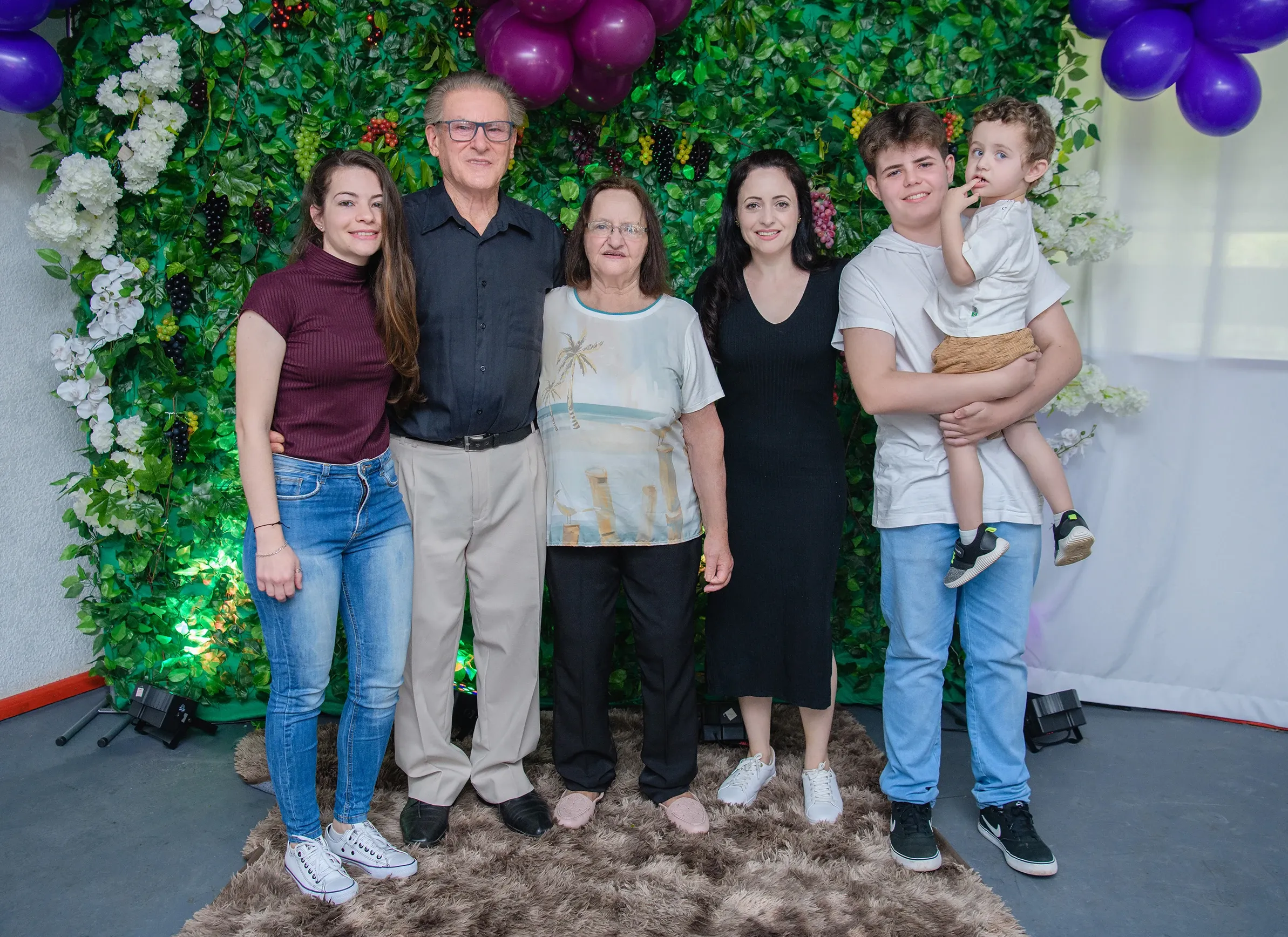 Grupo de seis pessoas de diferentes idades posando em frente a uma parede de folhagens e flores, com balões roxos no canto superior direito.