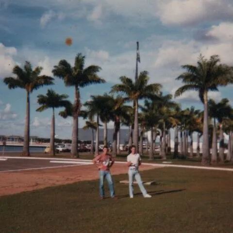 Dois homens parados em um gramado com palmeiras altas ao fundo, em um dia claro e ensolarado. (Original)