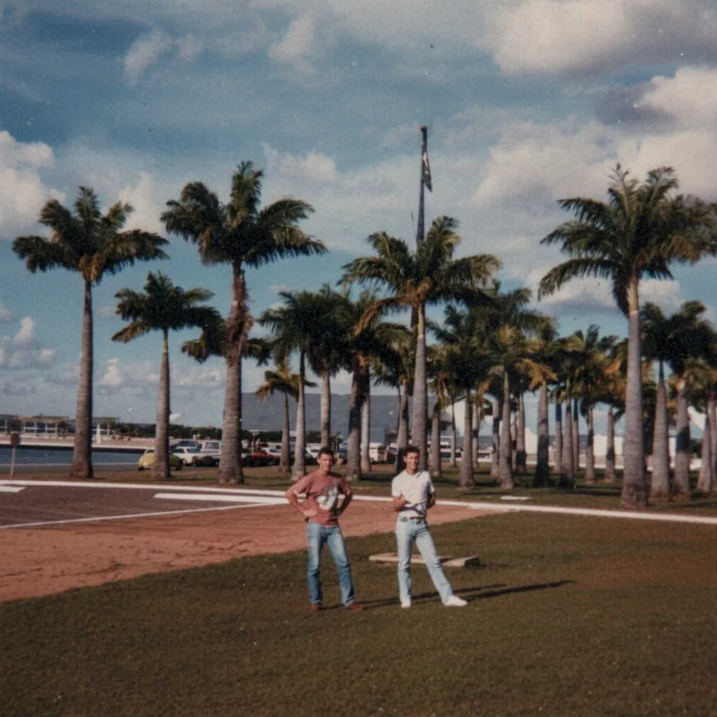 Dois homens parados em um gramado com palmeiras altas ao fundo, em um dia claro e ensolarado.