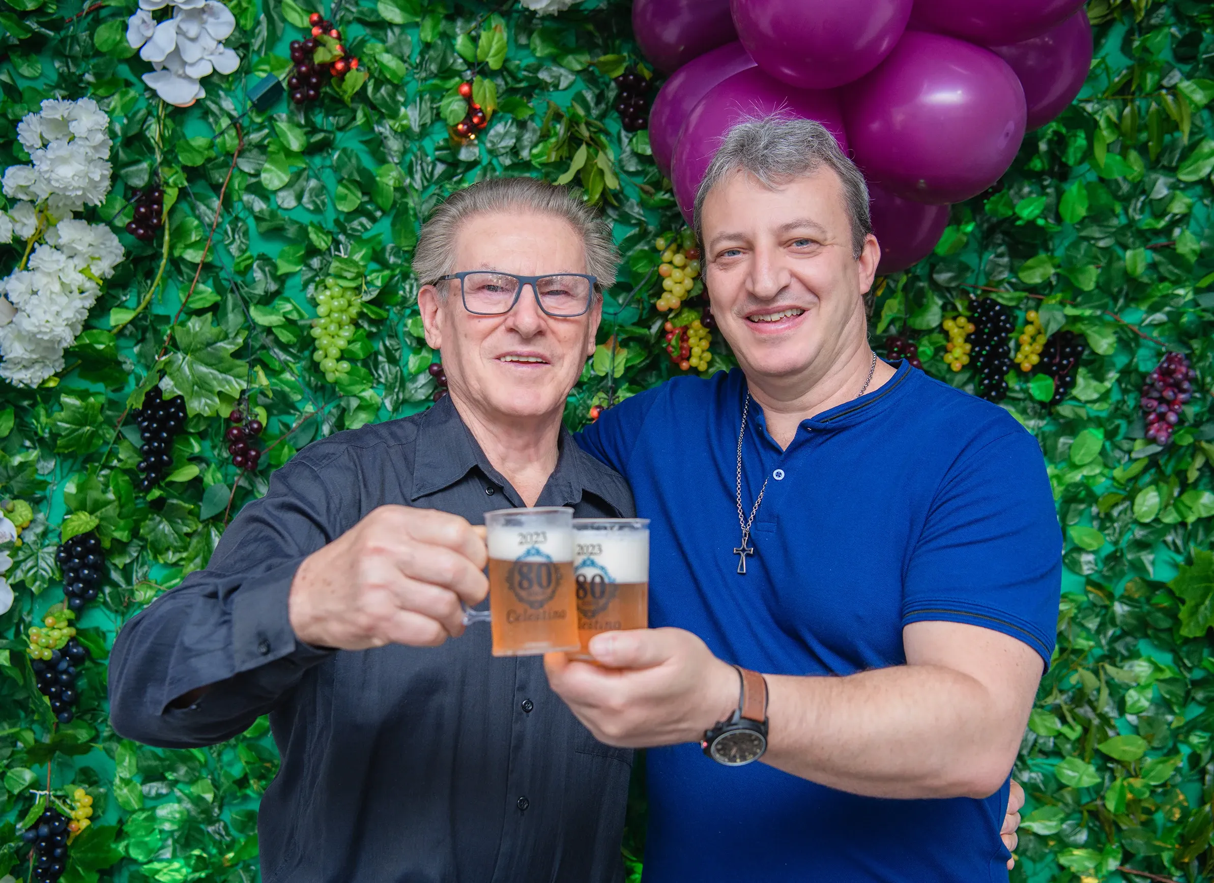 Dois homens sorridentes, um de camisa preta e óculos, o outro de camisa azul, erguem copos de cerveja personalizados com o número "80" e o nome "Celestino", com um fundo de parede de hera e balões roxos.