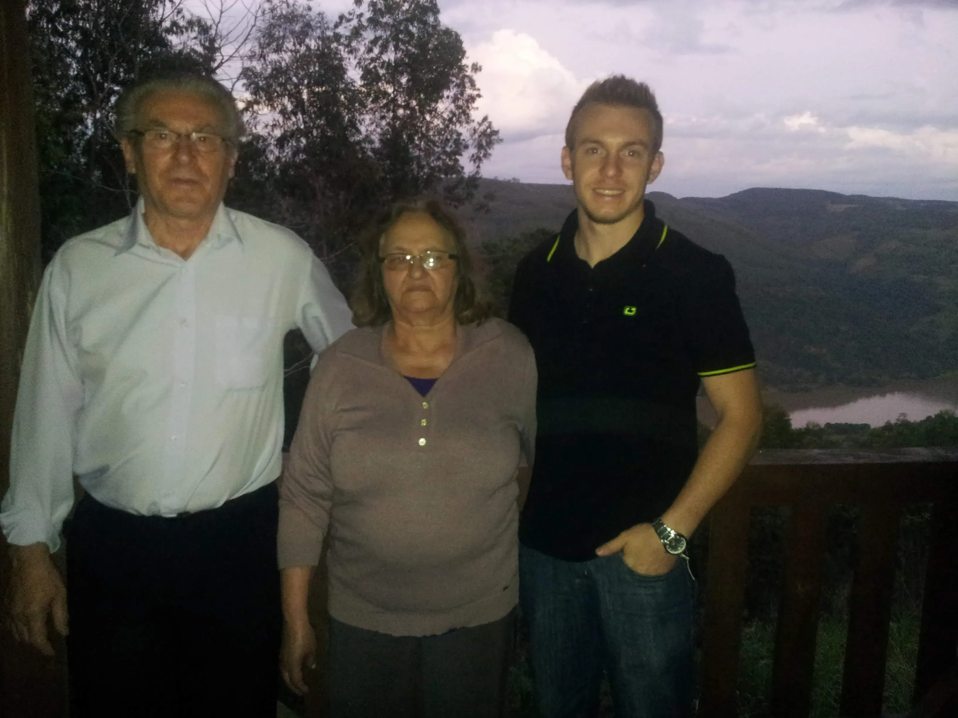 Homem idoso de óculos, mulher de meia-idade de óculos e jovem de camiseta polo preta posando lado a lado em uma varanda com vista para montanhas e um lago ao entardecer.