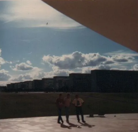 Três jovens caminham por uma área gramada com prédios residenciais ao fundo, sob um céu parcialmente nublado, com a parte superior direita da imagem obscurecida por uma estrutura angular. (Original)