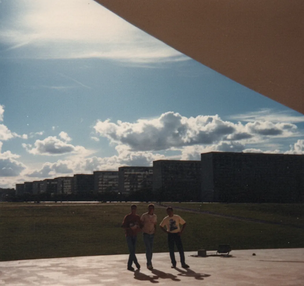 Três jovens caminham por uma área gramada com prédios residenciais ao fundo, sob um céu parcialmente nublado, com a parte superior direita da imagem obscurecida por uma estrutura angular.