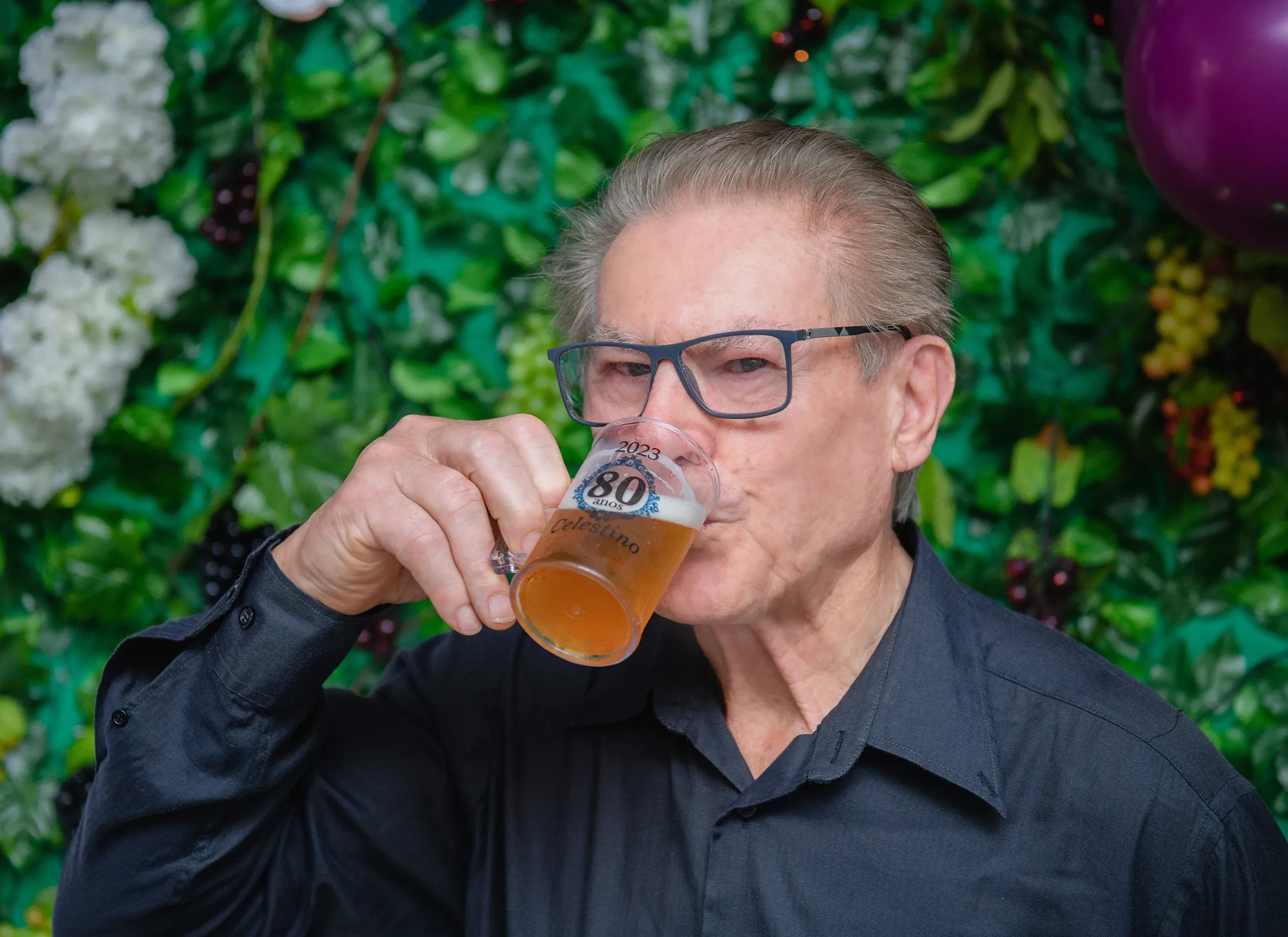 Homem idoso de óculos e camisa preta, bebendo em um copo que diz "2023 80 ANOS CELESTINO", com um fundo verde de folhagens.