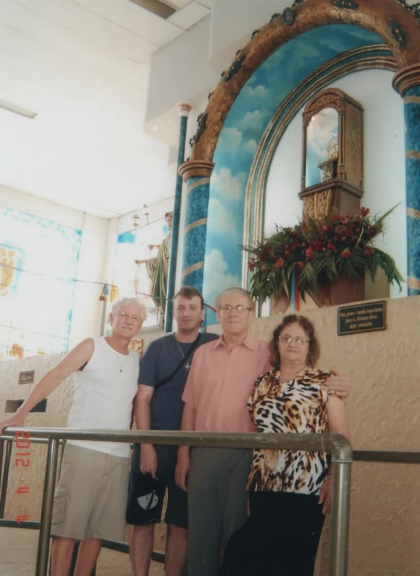Quatro pessoas, incluindo dois homens e duas mulheres, estão de pé na frente de um altar ornamentado com um arco azul pintado com nuvens brancas, dentro de um edifício bem iluminado.