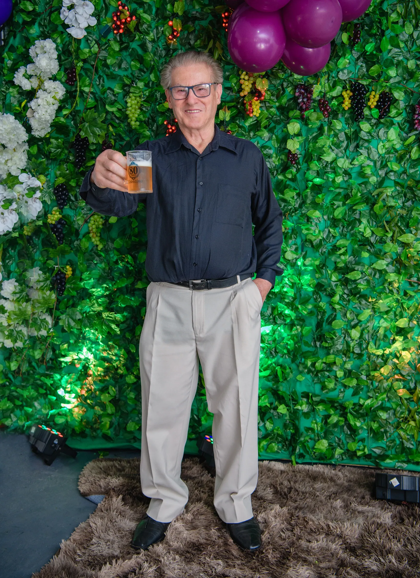 Homem idoso de óculos, camisa social escura e calças claras, em pé, sorrindo e segurando um copo de chopp, tendo ao fundo uma parede de folhas verdes decorada com flores brancas e uvas.