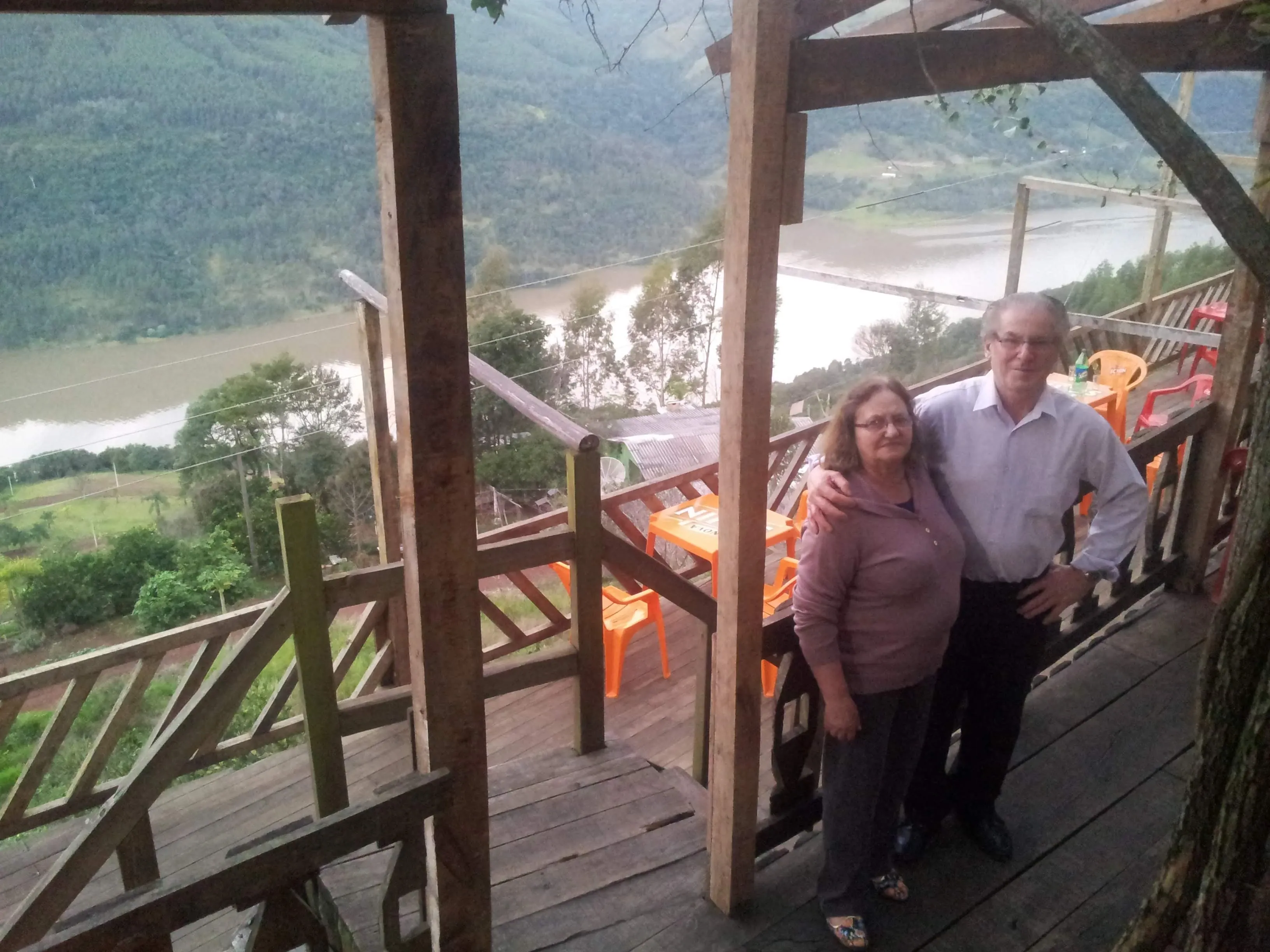 Casal de idosos abraçados em uma varanda de madeira com vista para um rio e montanhas.