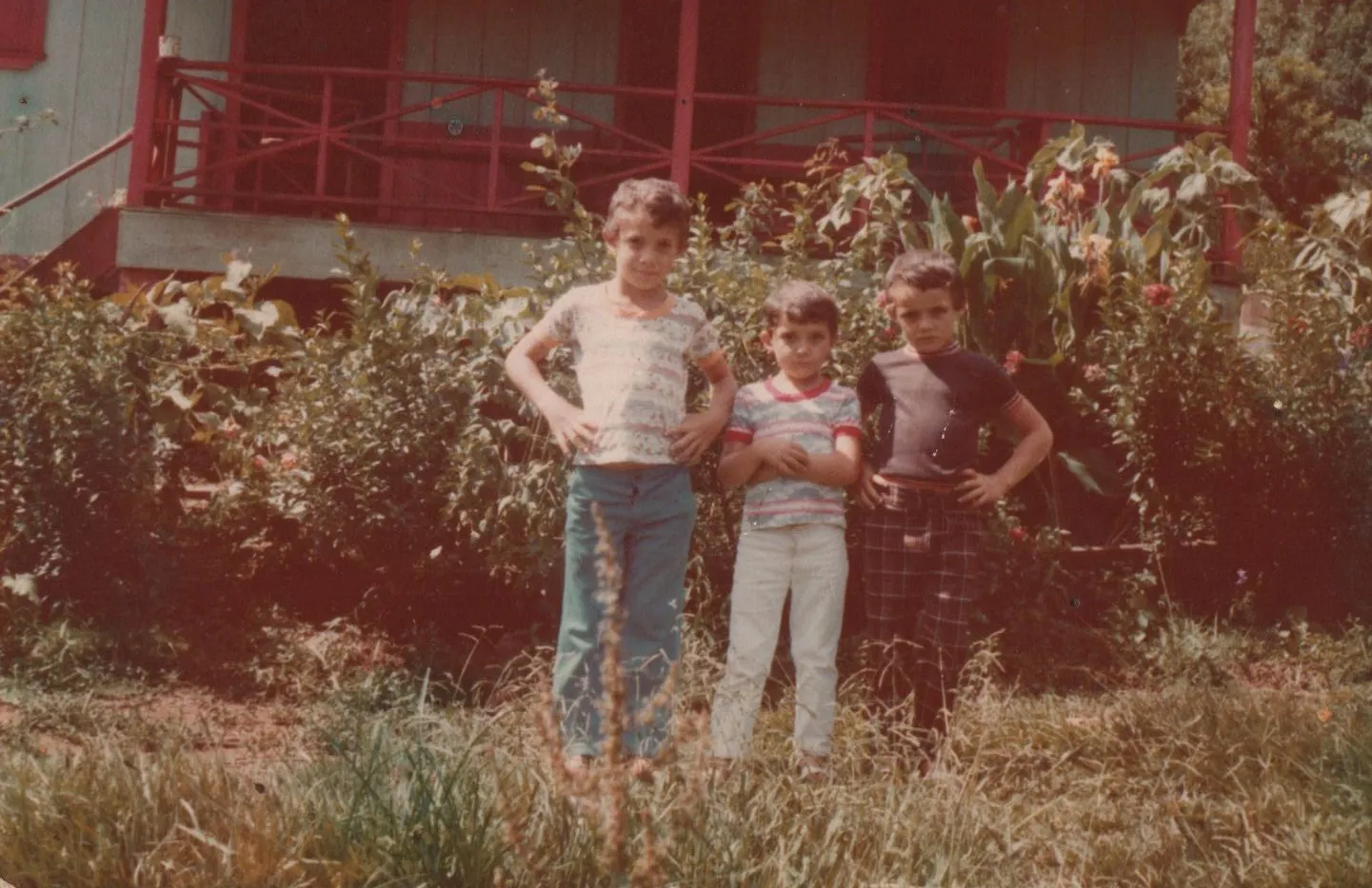Três meninos em idades variadas posam em frente a um alpendre vermelho e uma vegetação exuberante, olhando diretamente para a câmera. (Original)