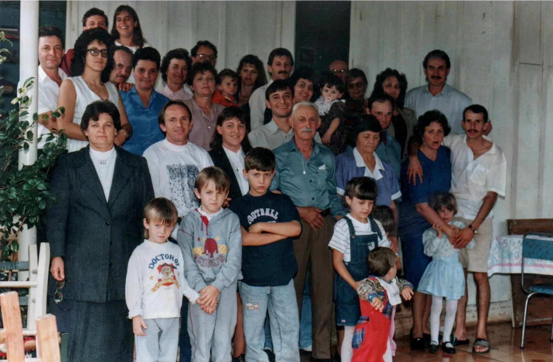 Uma grande família multi-geracional de homens, mulheres e crianças de várias idades está posando para uma foto em grupo na varanda da frente de uma casa.