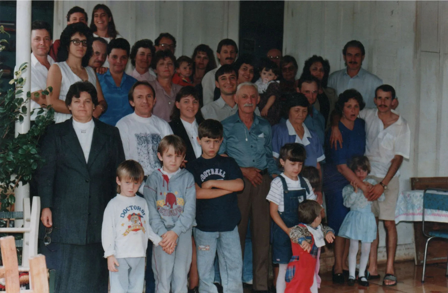 Uma grande família multi-geracional de homens, mulheres e crianças de várias idades está posando para uma foto em grupo na varanda da frente de uma casa. (Original)