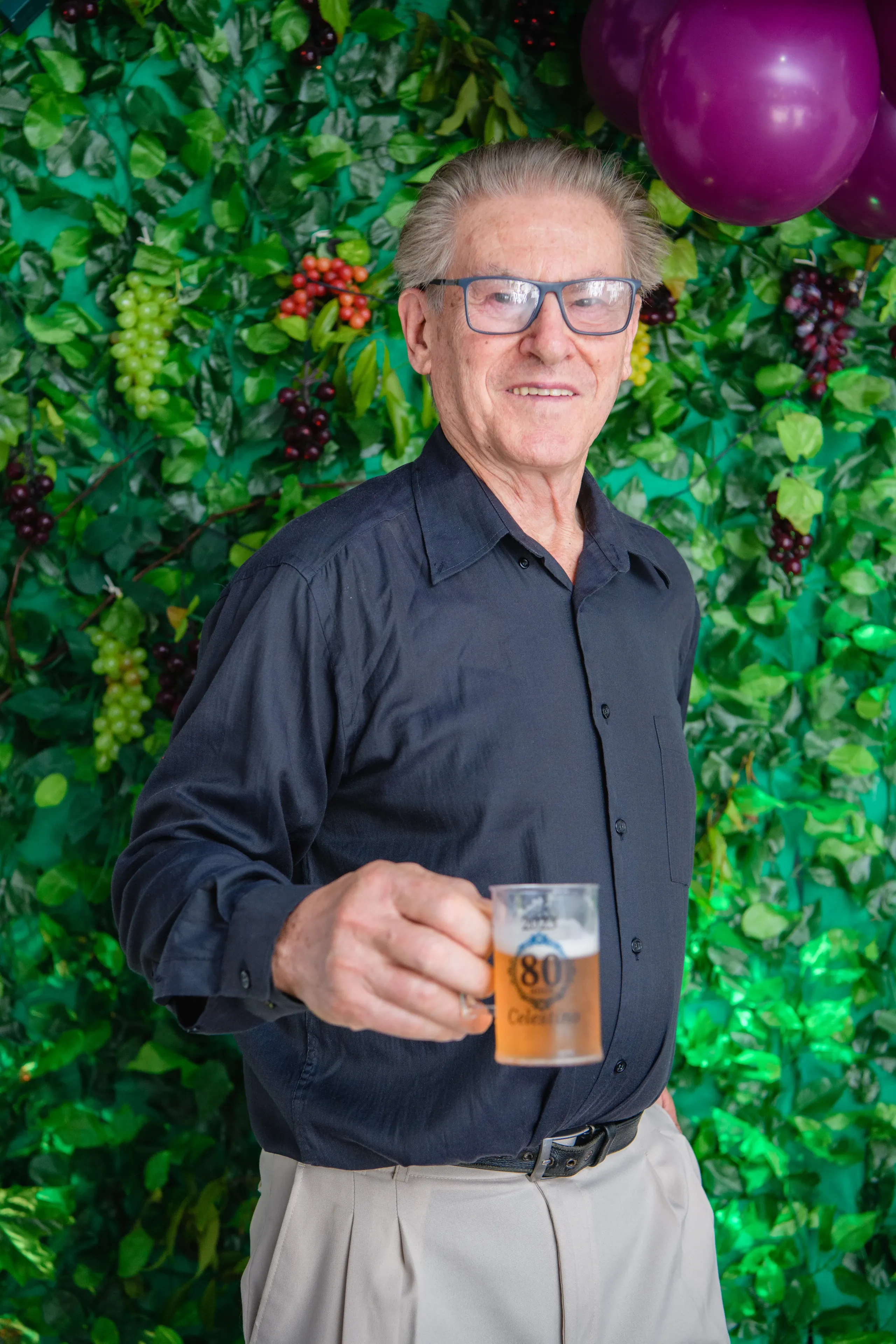 Homem idoso de óculos, camisa social escura e calças claras, em pé, sorrindo e segurando um copo de chopp, tendo ao fundo uma parede de folhas verdes decorada com flores brancas e uvas.