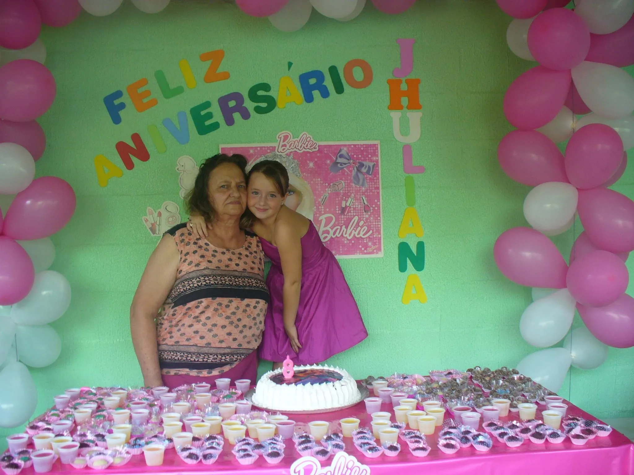 Uma mulher mais velha e uma criança em um vestido roxo se abraçam na frente de uma mesa de festa de aniversário com balões e doces.