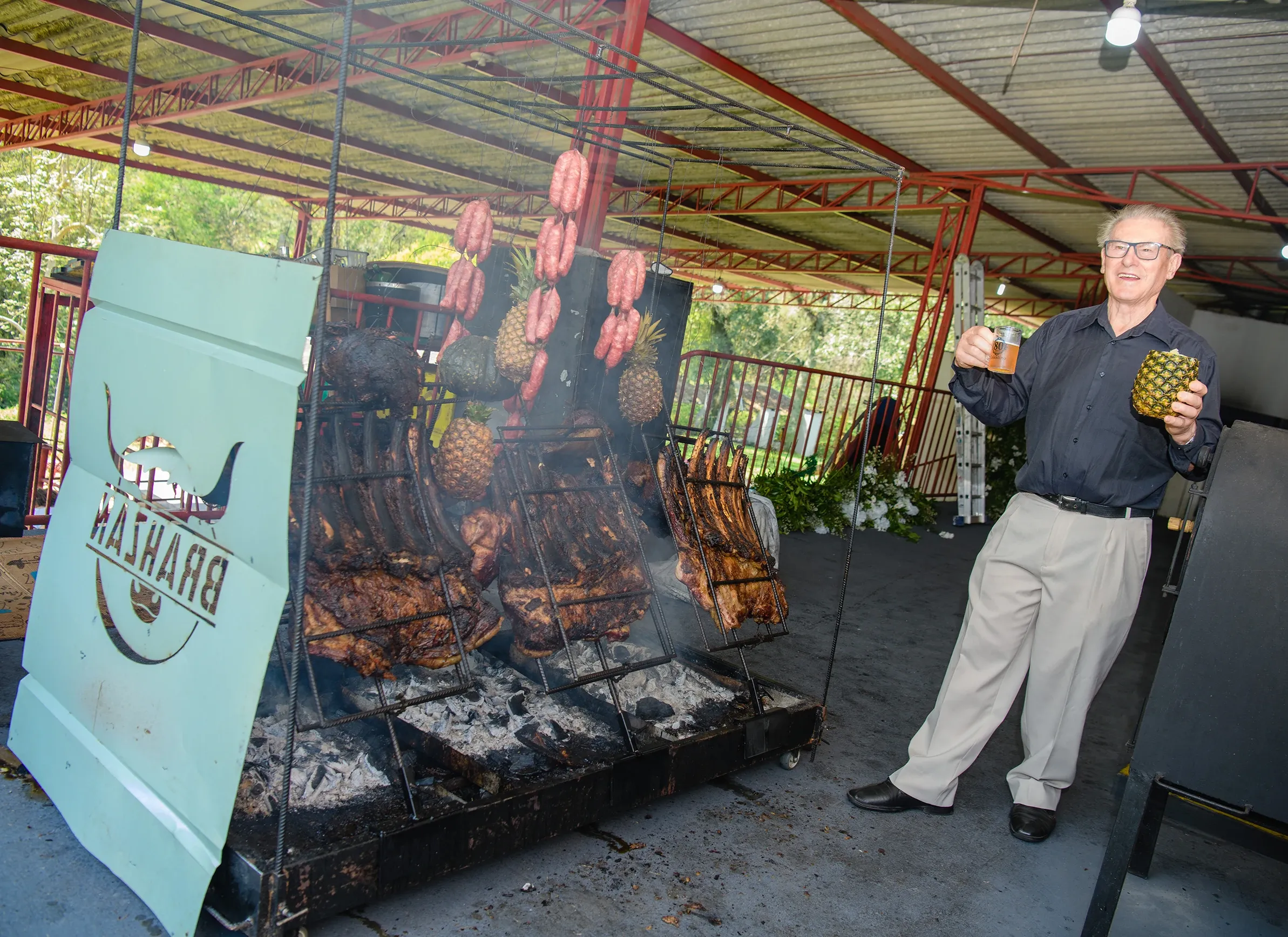 Um homem de óculos e camisa preta ergue um copo e um abacaxi ao lado de uma churrasqueira grande com vários espetos de carne e abacaxis, em um espaço aberto com cobertura metálica.