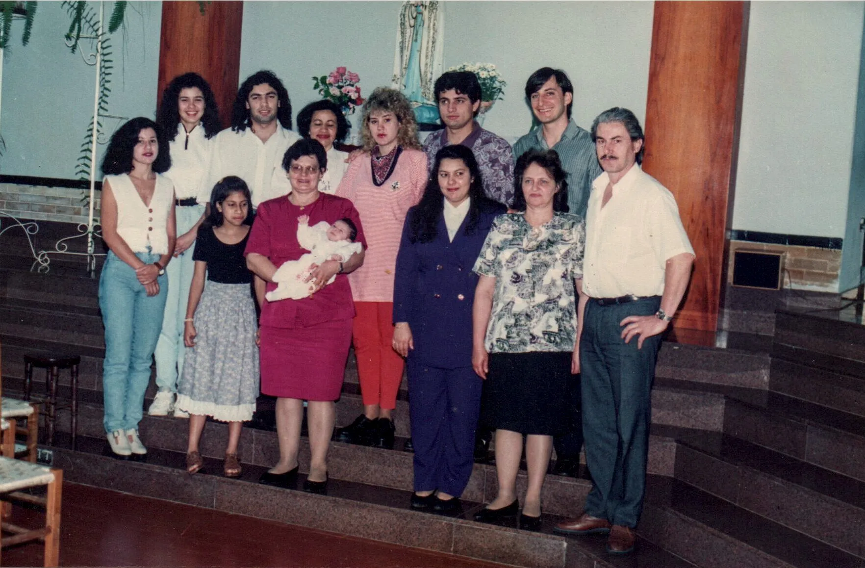Um grupo grande de pessoas está em uma escadaria, com mulheres de várias idades e crianças à esquerda e homens à direita. Uma mulher no centro segura um bebê nos braços.