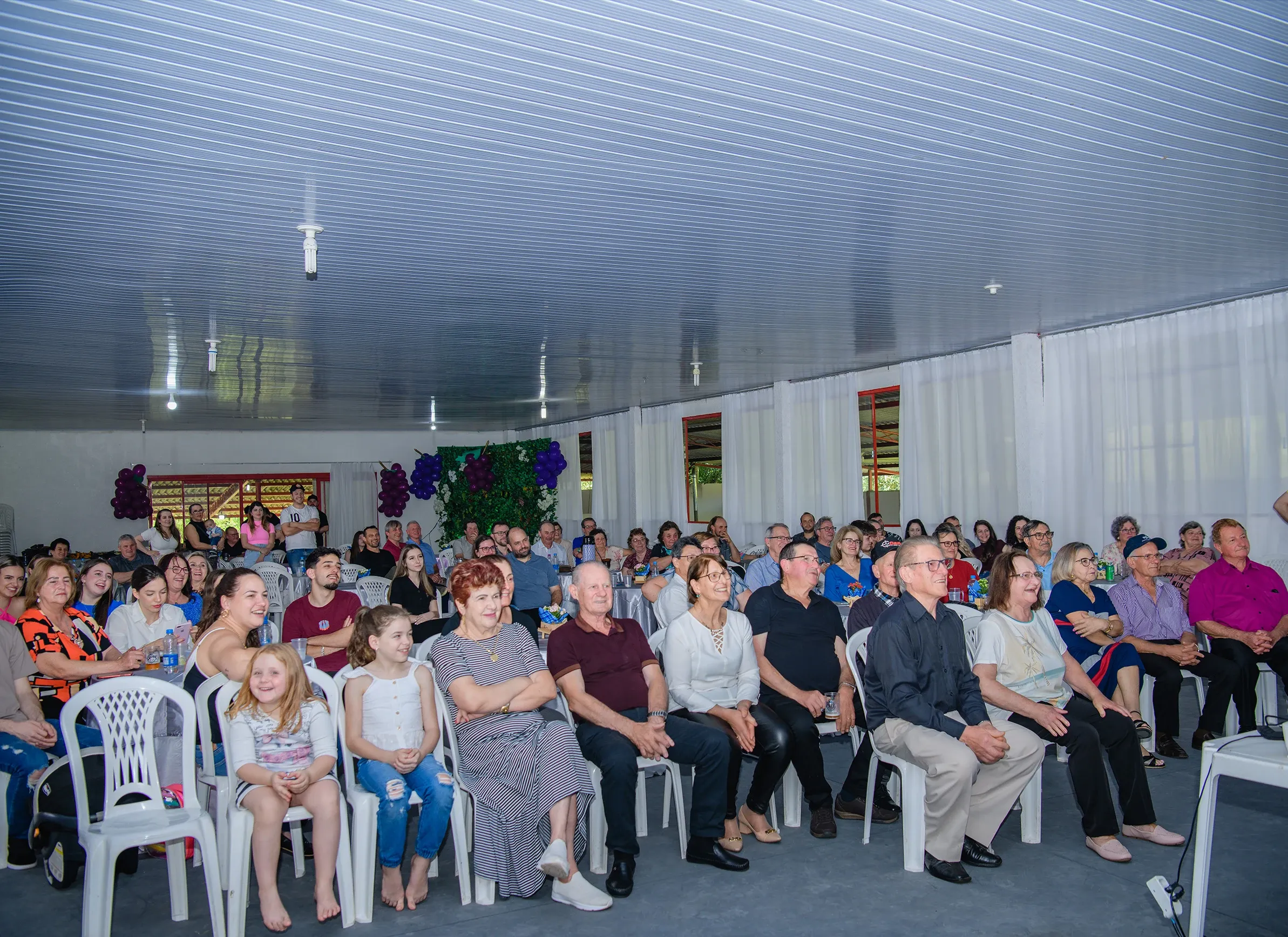 Uma grande plateia de pessoas de todas as idades, predominantemente adultas e idosas, sentada em cadeiras brancas e escuras em um auditório espaçoso com teto azul e cortinas brancas, olhando na mesma direção.