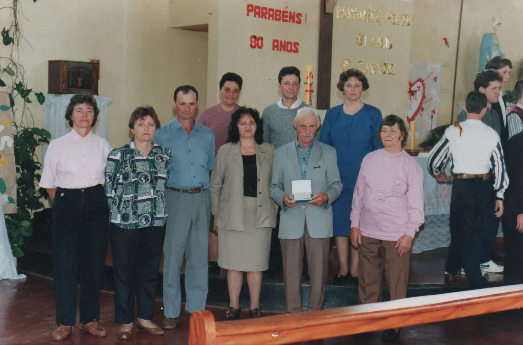 Um grupo de treze pessoas, algumas sorrindo, posa para uma foto em um evento formal, com um homem idoso no centro segurando uma pequena caixa ou placa, e uma mulher mais jovem à sua esquerda. (Original)
